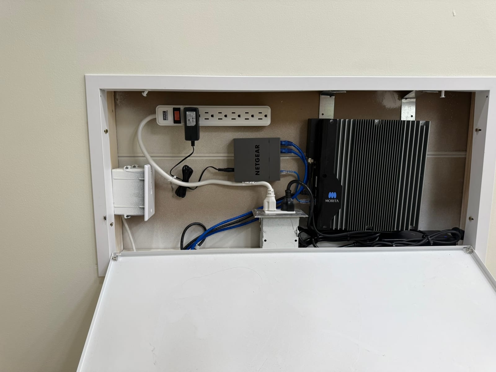 Open wall recess panel housing a power strip, Netgear network switch, and Morita control unit with cabling organized inside.